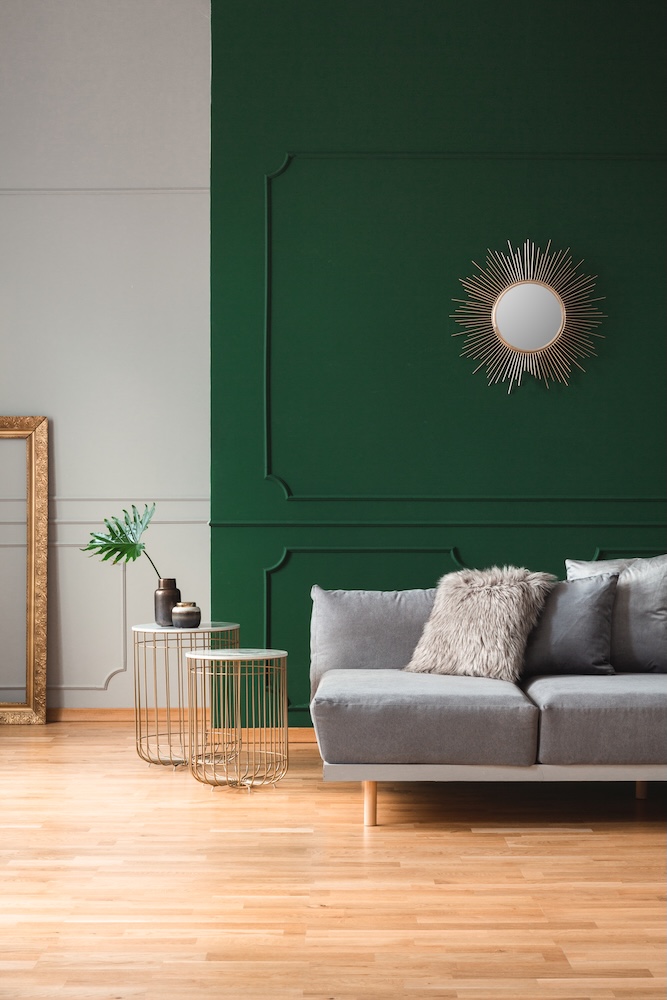 salon z szarą sofą przed ciemnozieloną ścianą z okrągłym lustrem ciemnozielona ściana za szarą kanapą w salonie z lustrem